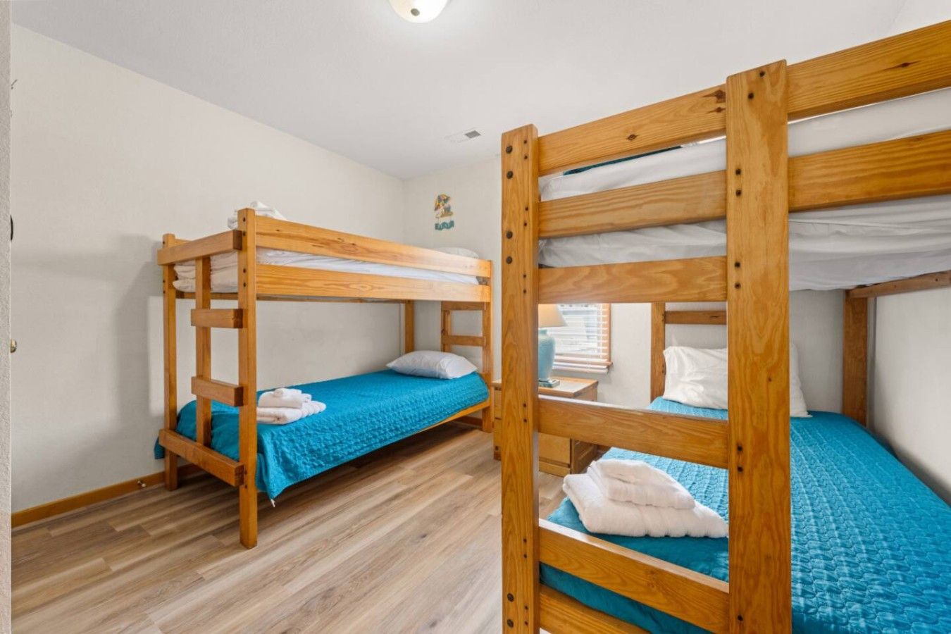 A room with two bunk beds, wooden flooring, and a small window with blinds.