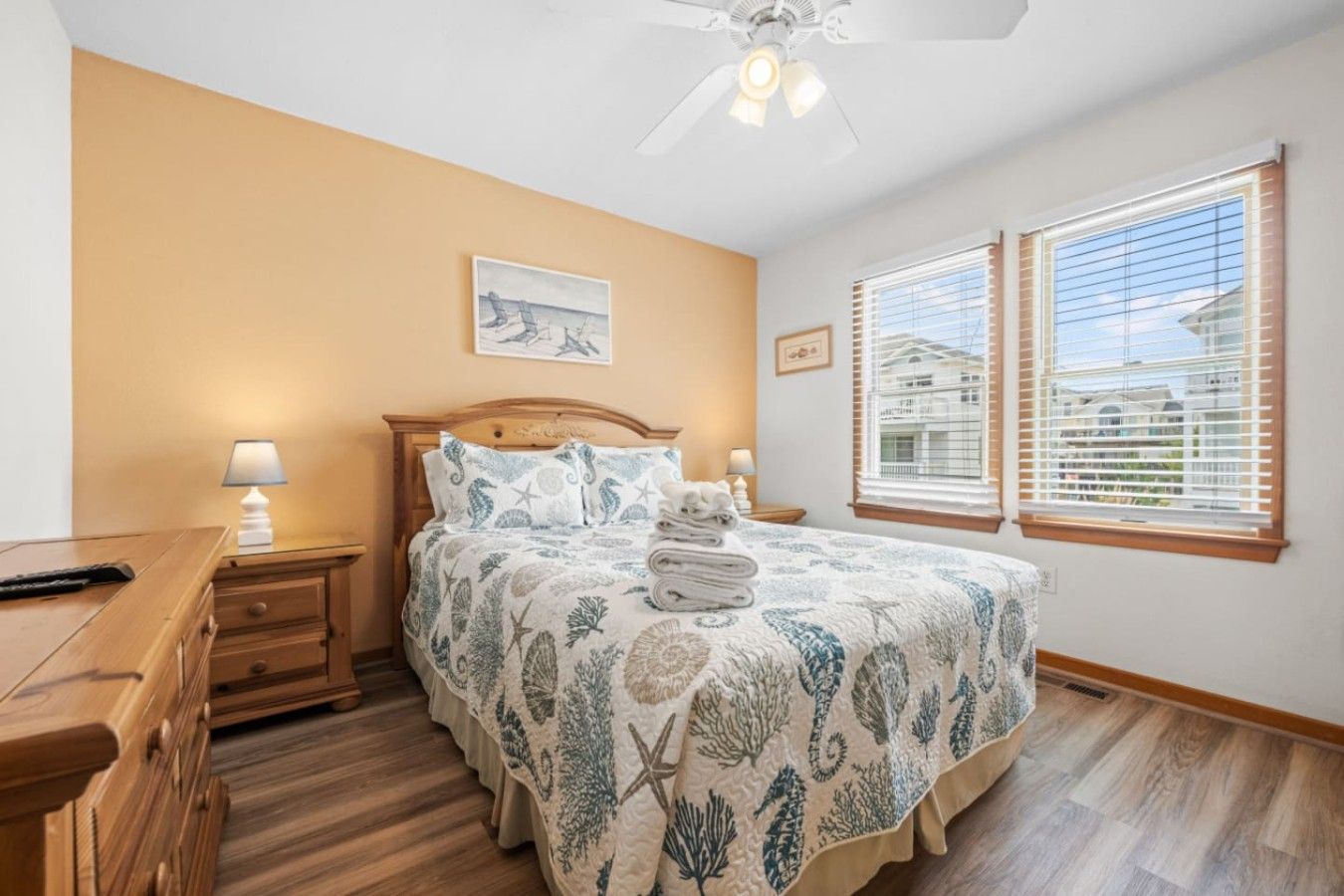 A bedroom with a bed, bedside lamps, a wooden nightstand, a ceiling fan, and two windows with blinds.
