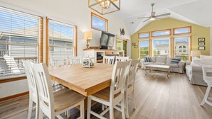 Un vaste espace de vie comprenant une table à manger, des chaises blanches, un téléviseur et un ventilateur de plafond.