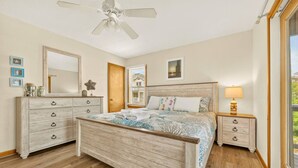Une chambre à coucher avec un lit en bois, une commode, une table de chevet, un ventilateur de plafond et une fenêtre donnant sur un beau paysage.