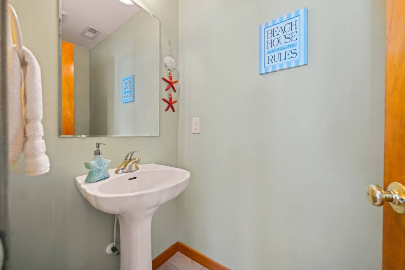 Ein Badezimmer mit einem weißen Waschbecken, einem Spiegel und einem Handtuchhalter. Ein Schild an der Wand zeigt die Aufschrift 'BEACH HOUSE RULES'.