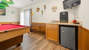 Une table de billard recouverte d’un feutre rouge, un meuble en bois, un réfrigérateur noir et un téléviseur fixé au mur.