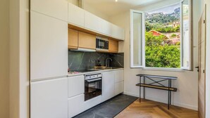 A modern kitchen with white cabinetry, a built-in oven, and a window with a view of greenery and buildings. - Elegant one-bedroom apartment with sea view at the gateway to Monaco (Beausoleil)