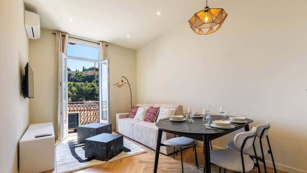 A modern living room with a dining area, a sofa with cushions, a coffee table, a TV, and a balcony with a view. - Elegant one-bedroom apartment with sea view at the gateway to Monaco (Beausoleil)