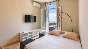 A modern bedroom with a bed, a TV, a white console, and a view of the city. - Elegant one-bedroom apartment with sea view at the gateway to Monaco (Beausoleil)