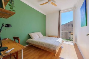 A bedroom with a bed, a desk, a chair, a ceiling fan, and a view of the outside.