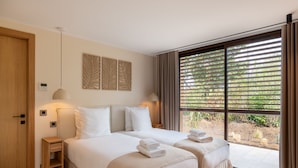 Ein modernes Hotelzimmer mit zwei Betten, einem großen Fenster mit Jalousien, einem Nachttisch und einer Wandleuchte.