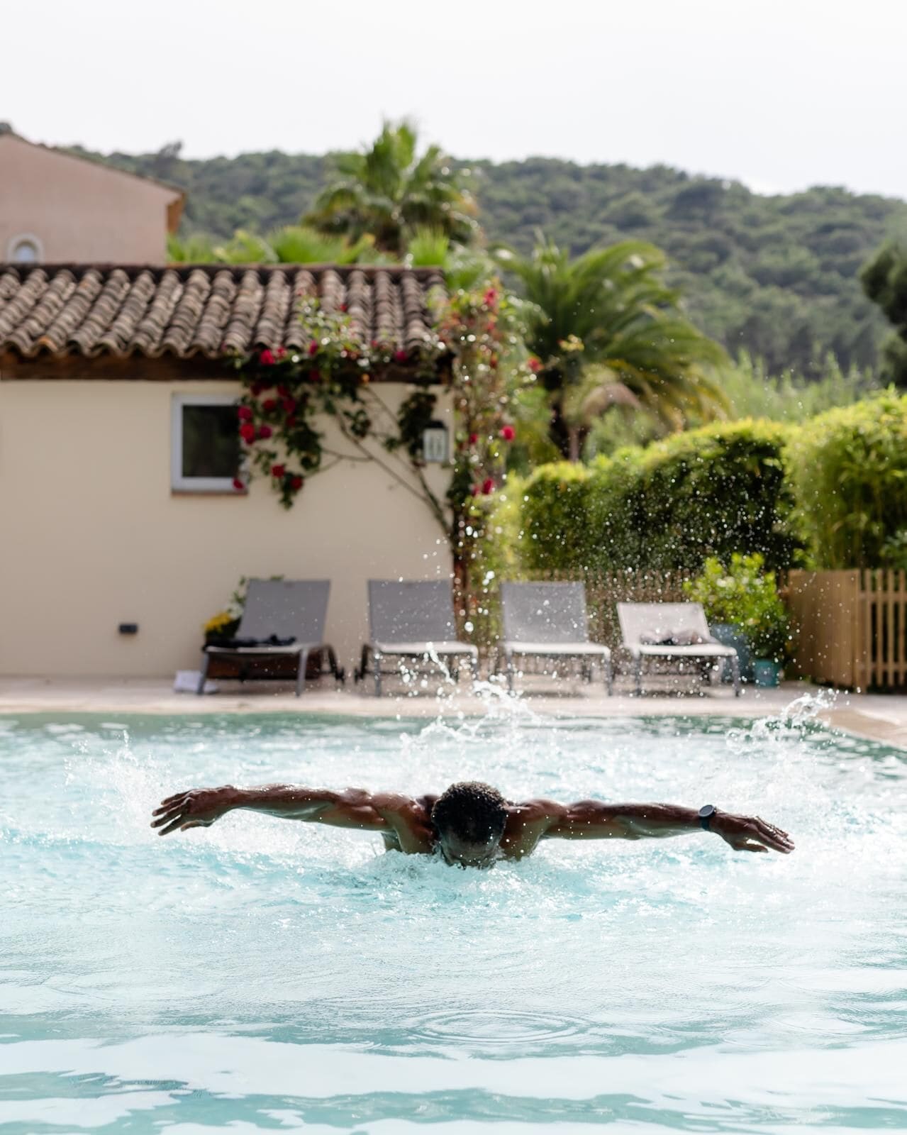 A swimming pool with a person swimming. There are lounge chairs and a building with a tiled roof in the background.