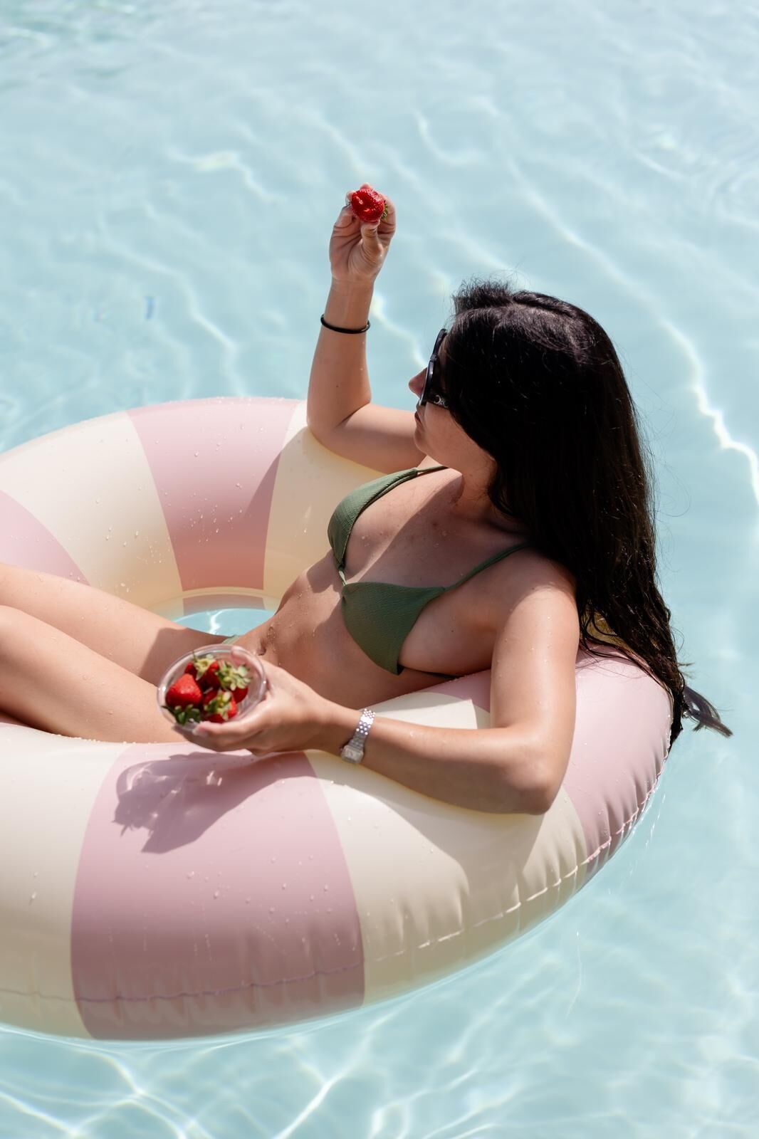 A person in a green bikini floating on a pink and white striped inflatable ring in a pool.