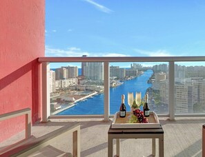A balcony with a view of a river and cityscape, a table with bottles and grapes, and a red wall.