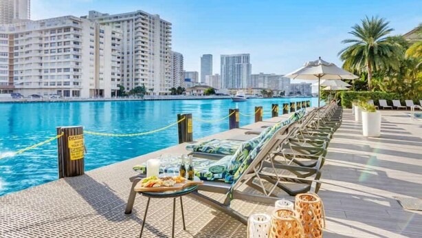 A poolside area with lounge chairs and a dining table, overlooking a body of water with buildings in the background. - Cozy Studio with Pool & Bay View Fitness Center (Hallandale Beach)