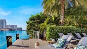A waterfront patio with lounge chairs, a small table, and a view of the marina and buildings. - Cozy Studio with Pool & Bay View Fitness Center (Hallandale Beach)