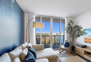 A modern living room with a sofa, a lamp, a TV, and a balcony with a view of the city. - 1BR Apartment with Balcony & amenities (Hallandale Beach)