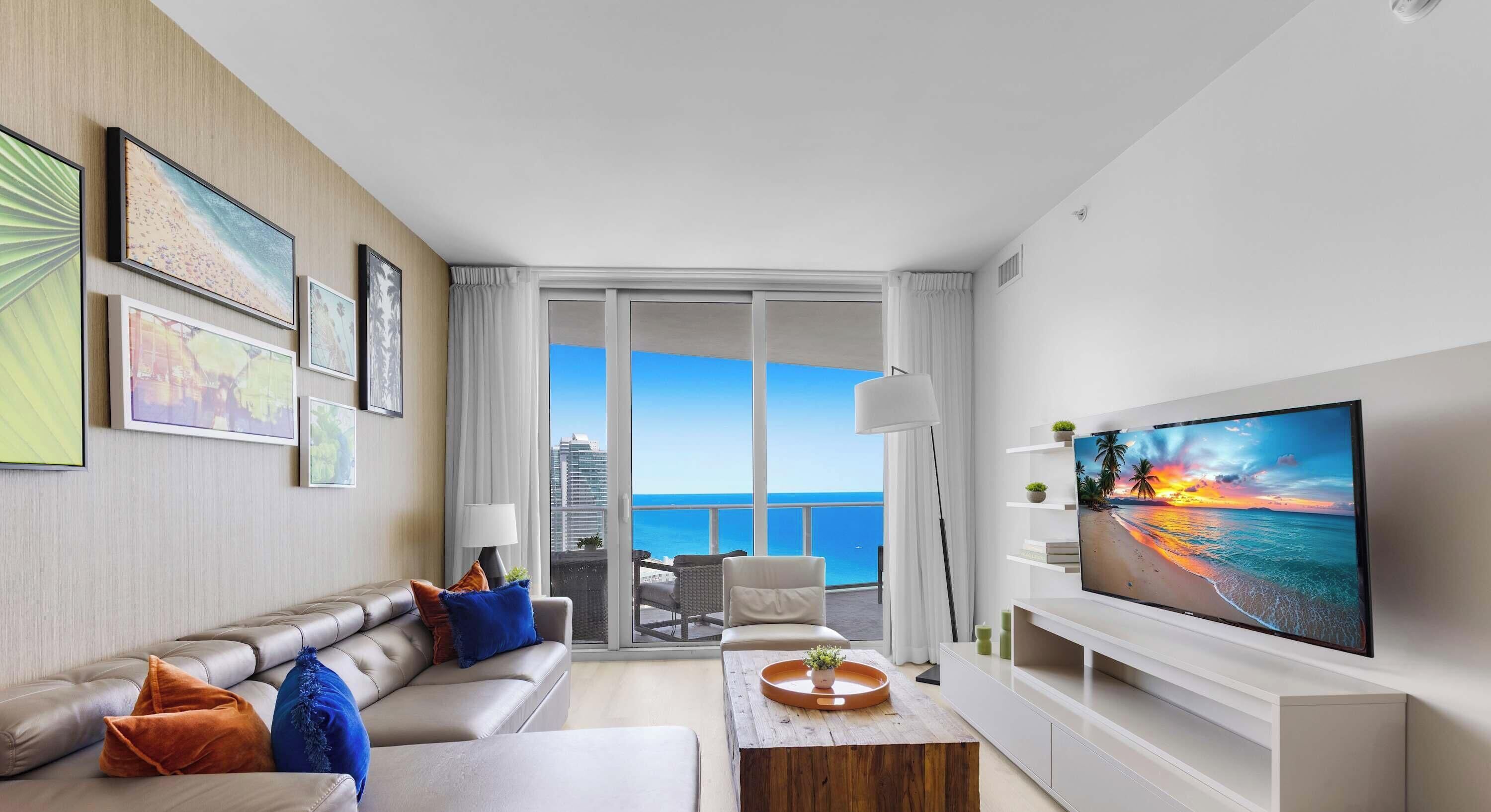 A modern living room with a large flat-screen TV, a sofa, a coffee table, and a balcony with a city view.