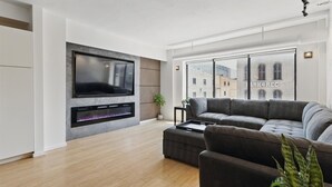 A modern living room with a flat-screen TV, a fireplace, a grey sectional sofa, and a view of a building with 'ARKGREC' on the window. - Modern Flat in the Heart of Indianapolis (Indianapolis)