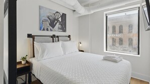 A bedroom with a bed, a nightstand, a lamp, a painting, and a window with a view of a building. - Modern Flat in the Heart of Indianapolis (Indianapolis)