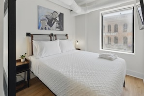 A bedroom with a bed, a nightstand, a lamp, a painting, and a window with a view of a building.