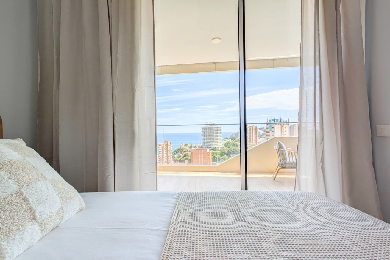 Een slaapkamer met een bed, uitzicht op een stadsbeeld door grote ramen en een balkon met een stoel.