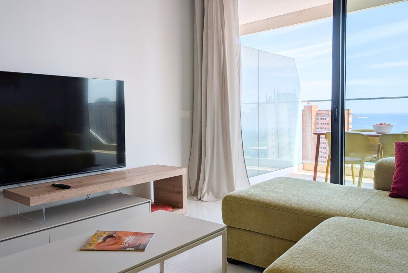 Un salon moderne doté d’un téléviseur à écran plat, d’une table basse avec des magazines, d’un canapé et d’un balcon offrant une vue sur la ville et la mer.
