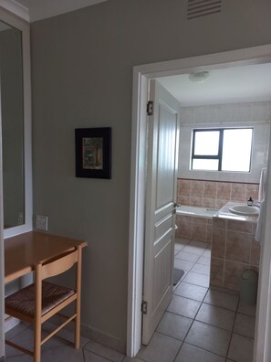 Ein Zimmer mit Holztisch und Stuhl, einem gerahmten Bild an der Wand sowie einem Badezimmer mit Fenster und Waschbecken.