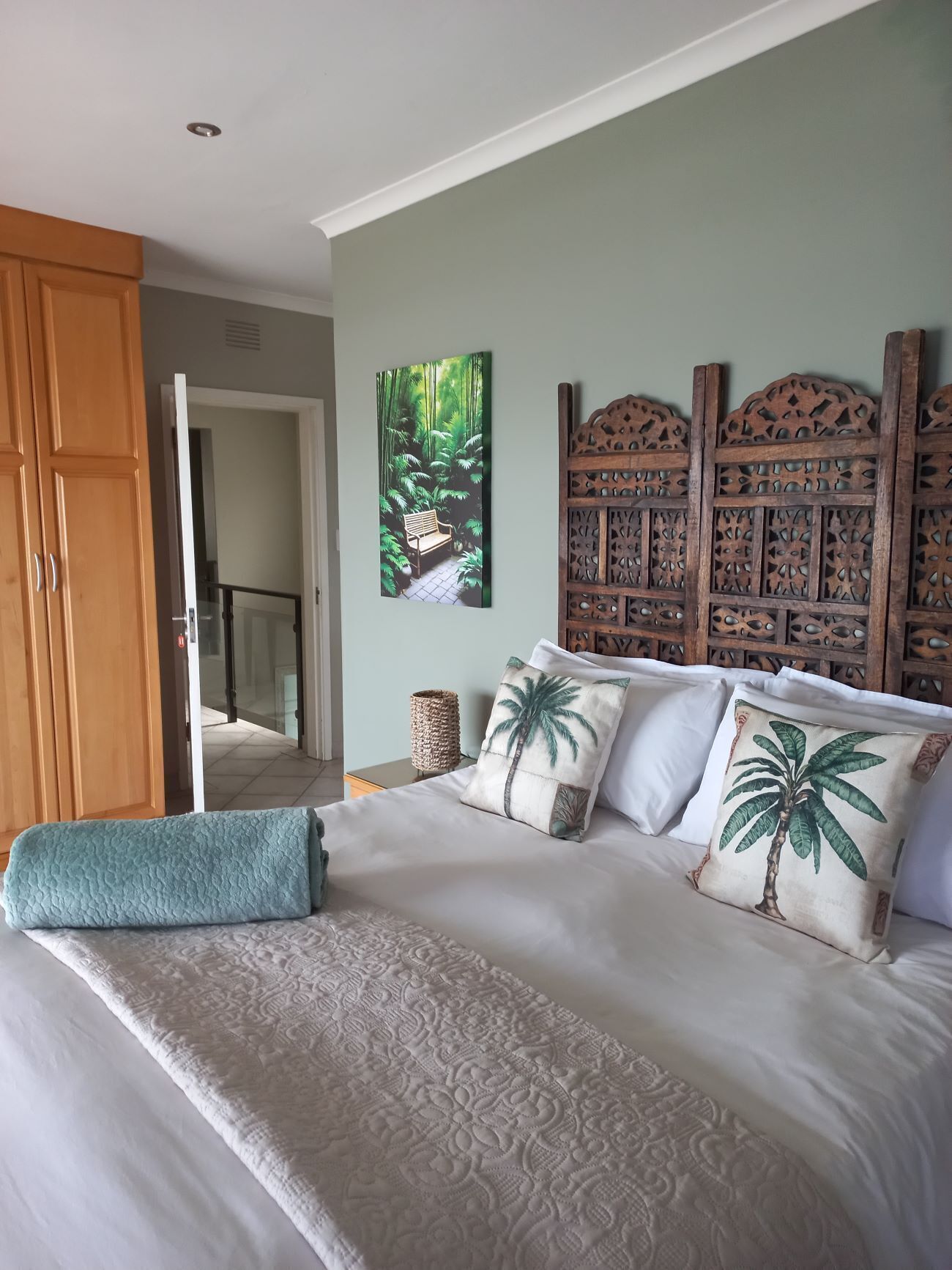 A bedroom with a bed, a wooden headboard, a painting of a forest, and a door leading to another room.