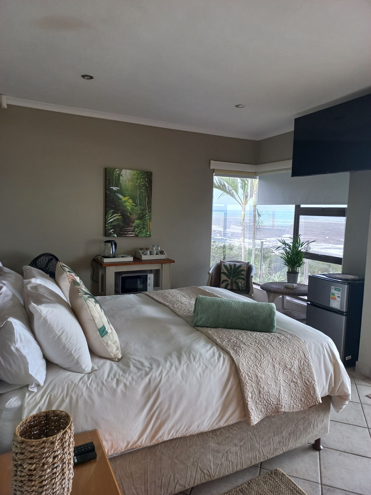 A bedroom with a large bed, a small table with a radio, a window with a view of palm trees, and a refrigerator.