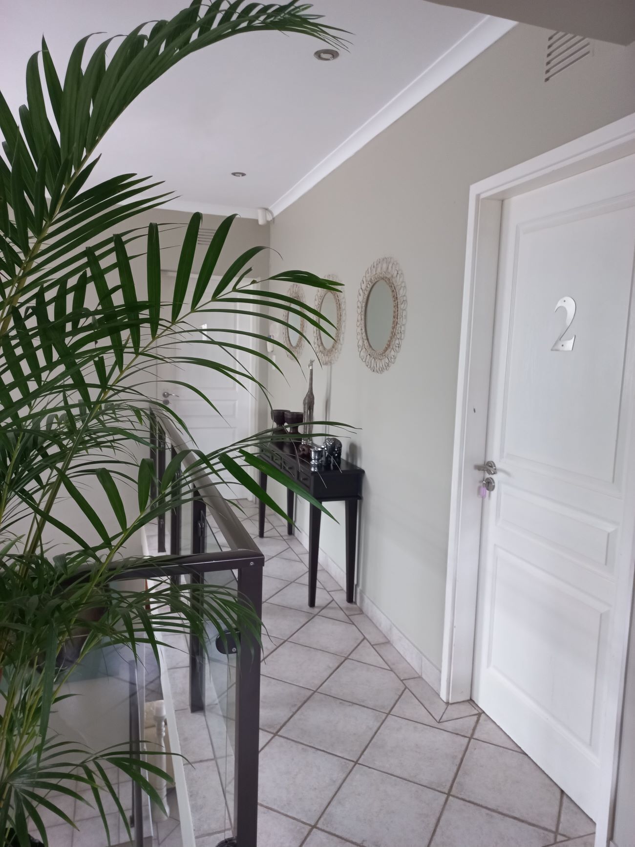 A hallway with a glass railing, a potted plant, a black console table, decorative mirrors, and a white door with the number 2.