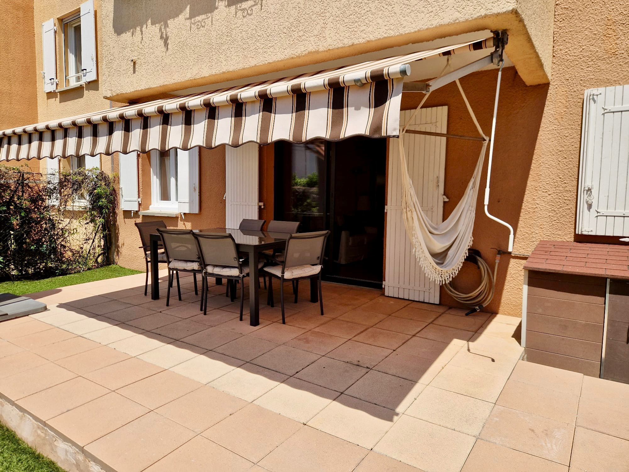 Une terrasse extérieure avec une table et des chaises, un hamac et un espace abrité doté d’un auvent à rayures.