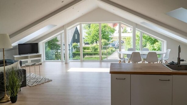 A modern loft apartment with a high ceiling, a dining area with a table and chairs, a kitchen with a sink, and a large sliding glass door leading to a garden. - 6 person modern apartment with style and comfort (Lejre)