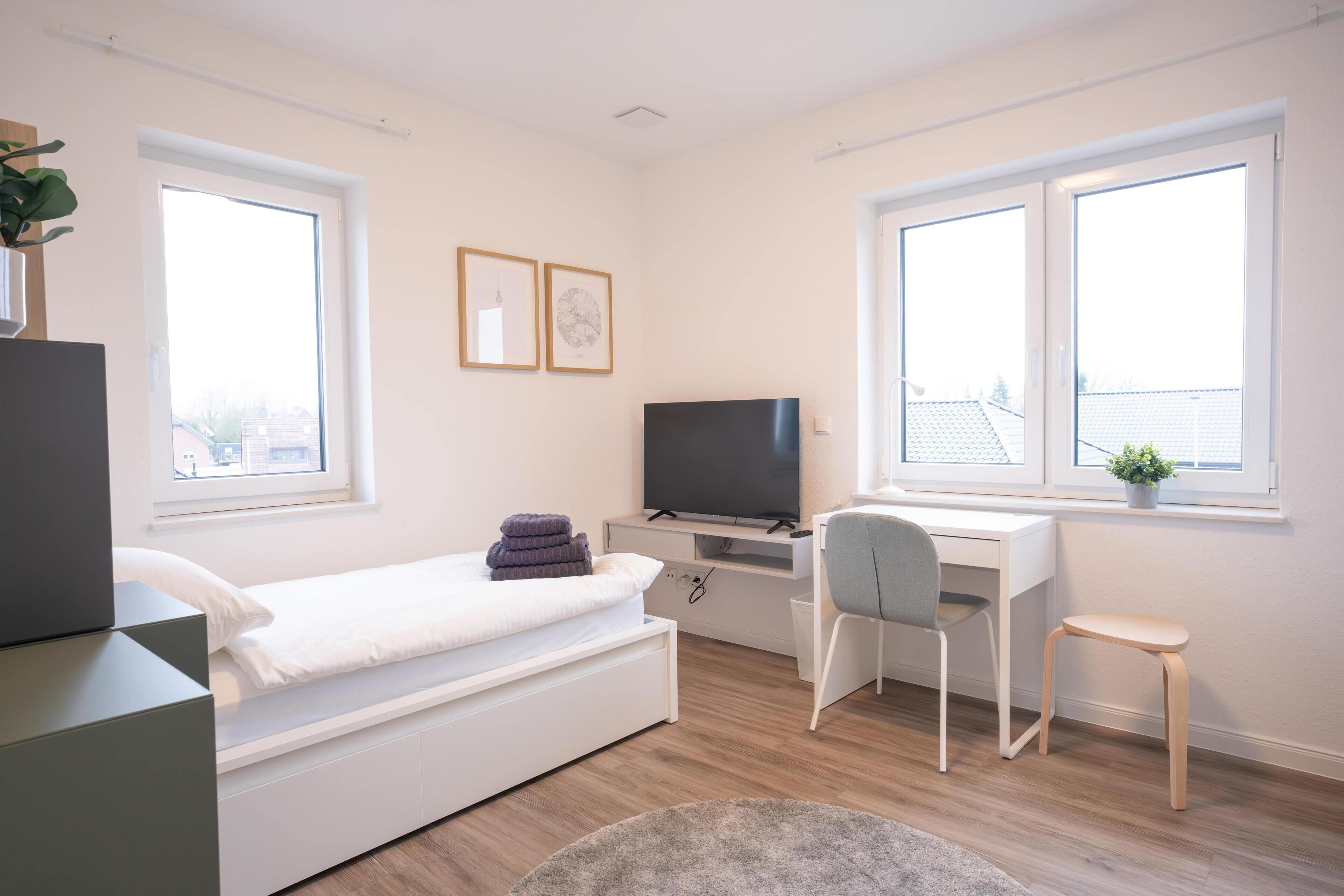 Une chambre moderne avec un lit, un bureau, une chaise, une télévision et de grandes fenêtres.