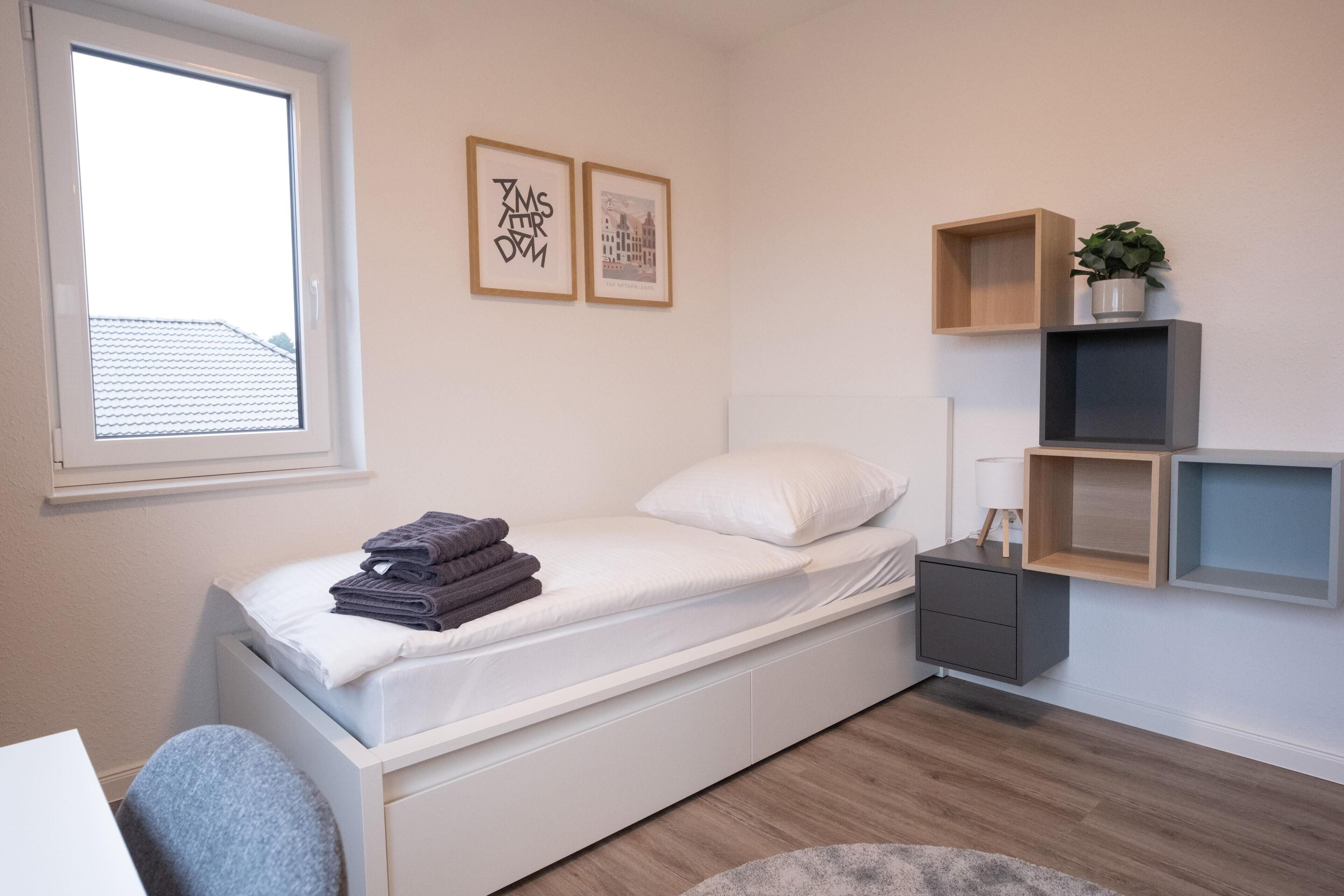 A small, neatly arranged bedroom with a single bed, a bedside table, a chair, a window, and wall-mounted shelves with a plant.