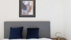 A neatly made bed with two dark pillows, a framed abstract painting above, and a bedside table with a globe lamp.