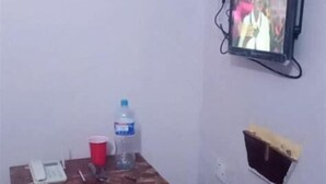 Een kleine kamer met een houten tafel, een rode emmer, een waterfles en een televisie die aan de muur hangt.