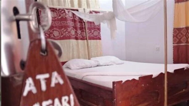 Een bed met een witte matras en een houten hoofdbord, een tas met de tekst 'ATA SAFARI LODGE' en een gordijn met een rood-beige patroon.