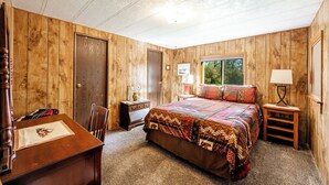 A bedroom with wooden walls, a bed with a colorful quilt, a desk with a chair, and a window with blinds.