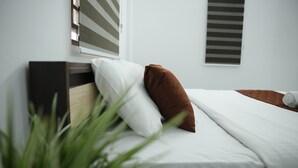 A neatly made bed with white and brown bedding, a wooden headboard, and a window with blinds. - Goldsuite (Kanayannur)