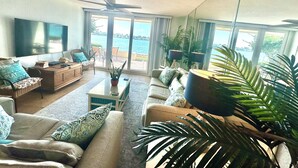 A spacious living room with a large TV, comfortable seating, and a view of the ocean through large windows.