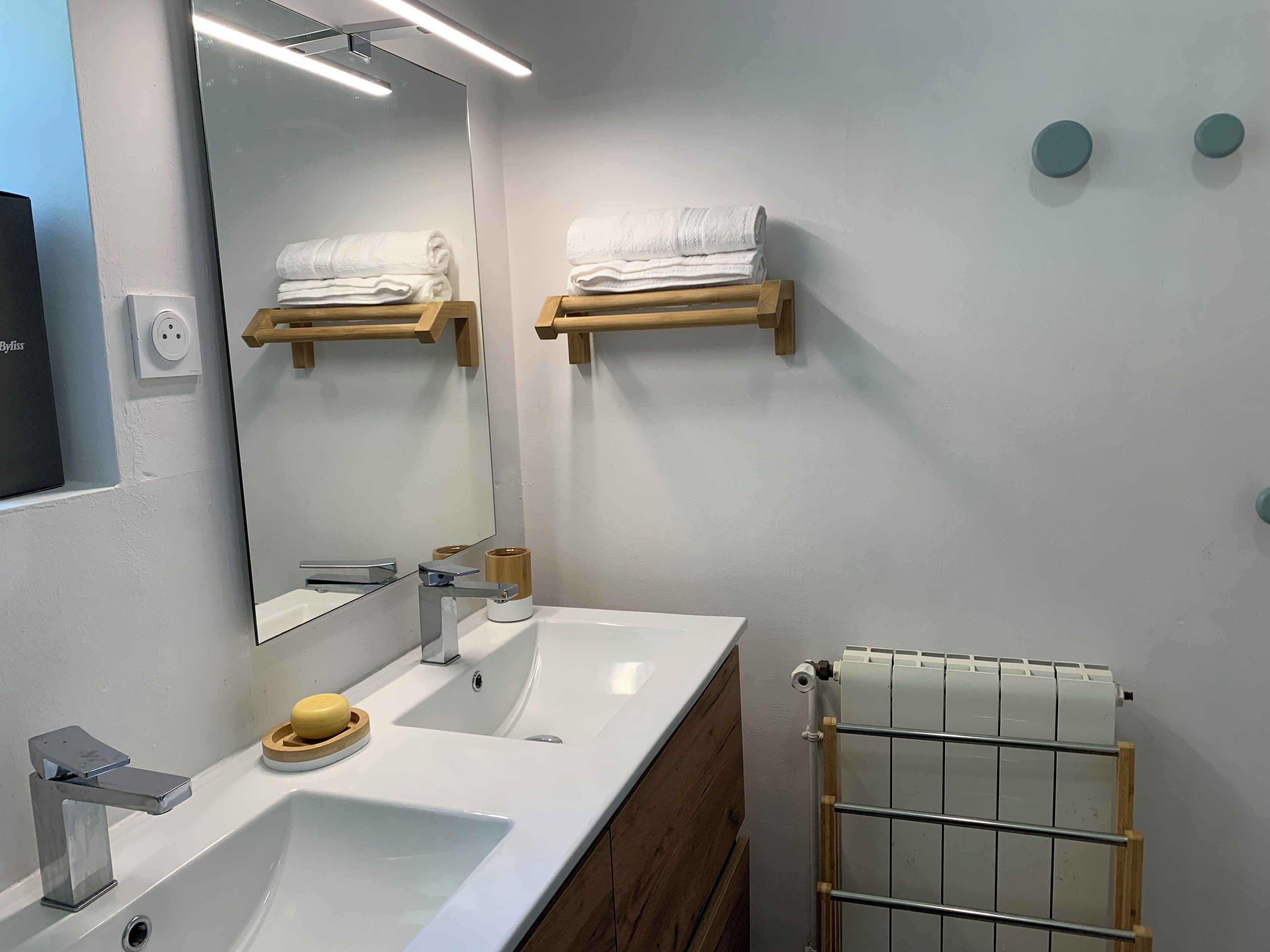 Uma casa de banho moderna com um espelho grande, duas pias, suportes de toalhas em madeira, um aquecedor de toalhas e um saboneteira.