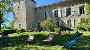 Un edificio de piedra con una torre, un jardín con césped y árboles, y tres sillones de descanso.
