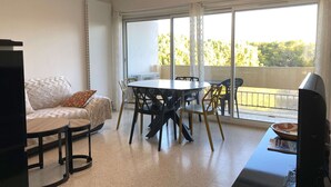 Un salon moderne avec une table à manger ronde, des chaises noires, un canapé blanc et un téléviseur. De grandes fenêtres offrent une vue sur l’extérieur.
