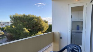 Un balcon avec une table et des chaises, une porte donnant sur un coin cuisine, et une vue sur les arbres et les bâtiments en arrière-plan.