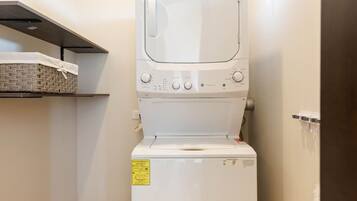 A compact, white front-loading washing machine in a small laundry room with a shelf above it.