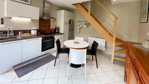 Une cuisine moderne avec des armoires blanches, une hotte en acier inoxydable et un four encastré. On y trouve un coin repas avec une table ronde et deux chaises. Un escalier en bois mène à l’étage supérieur.