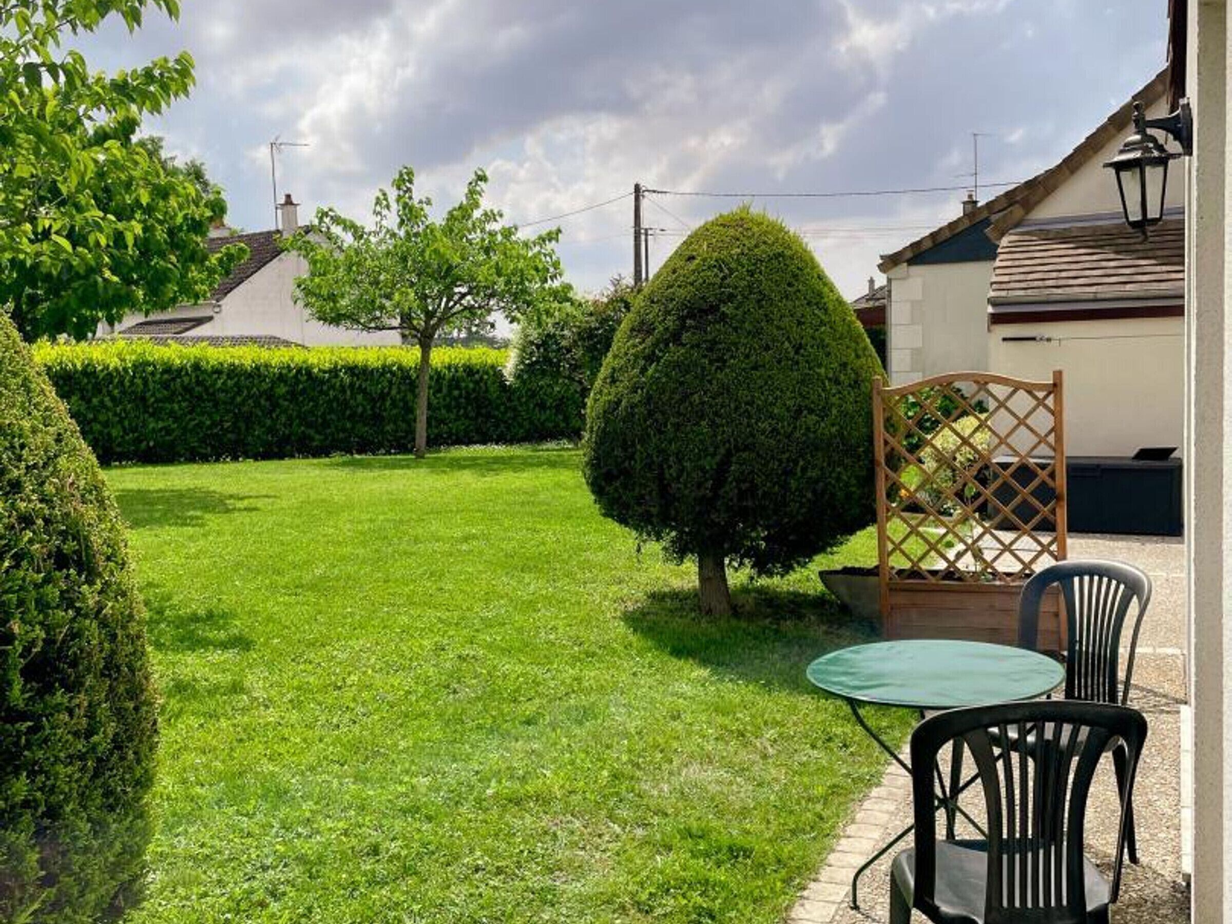 Un jardin bien entretenu, avec une pelouse soigneusement taillée, une table ronde entourée de chaises et une structure en treillis de bois.