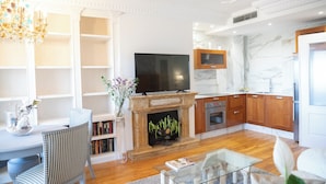 Uma sala de estar com lareira, televisão, área de jantar com mesa e cadeiras, e cozinha com armários de madeira e painel de mármore.