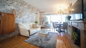 Uma sala de estar com um sofá branco, uma mesa de centro de vidro, piso de madeira, papel de parede estampado e um lustre.