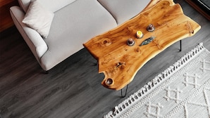 Uma sala de estar moderna com um sofá cinzento, uma mesa de centro de madeira com objetos decorativos e uma janela grande com cortinas.
