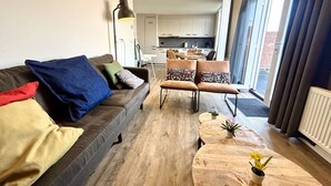 A modern living room with a sofa, armchairs, and a coffee table. There is a kitchen in the background and a sliding glass door leading to an outdoor area.