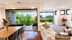 A modern living room with a wooden dining table, a sofa, and a balcony with a view of greenery. - Melbourne Beachfront Family Home with panoramic view (Black Rock)