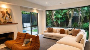 A living room with a fireplace, a sectional sofa, a brown armchair, and a large sliding glass door leading to a backyard with tropical plants. - Melbourne Beachfront Family Home with panoramic view (Black Rock)